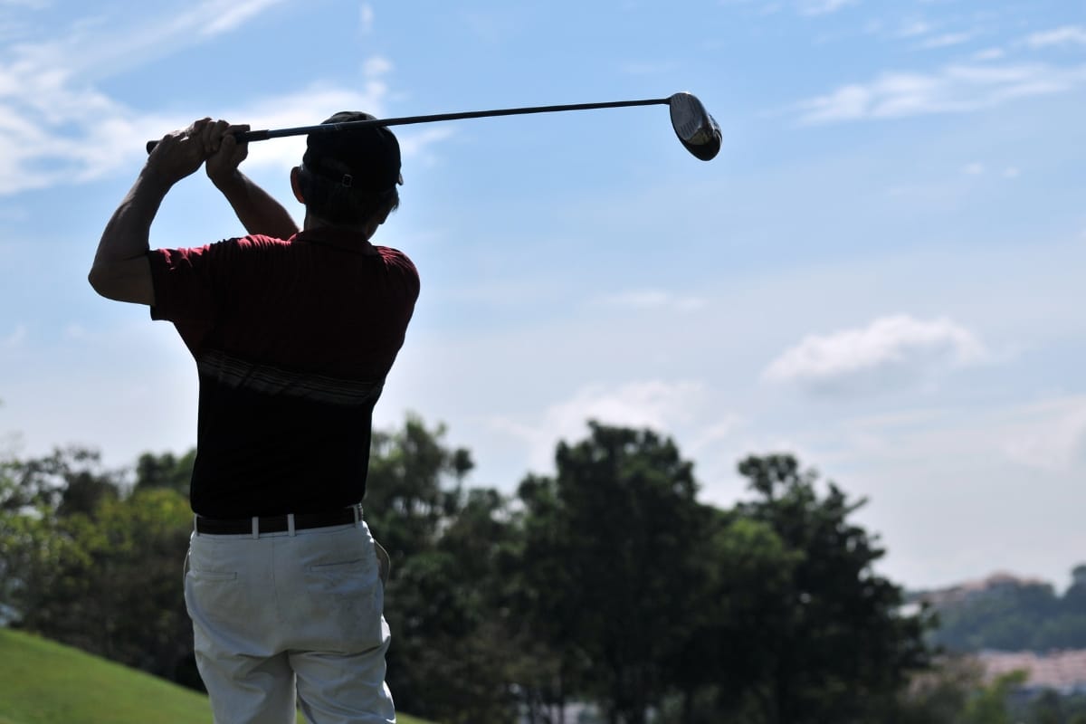 A golfer mid-swing, showcasing impressive golf performance on a sunny day with trees in the background.