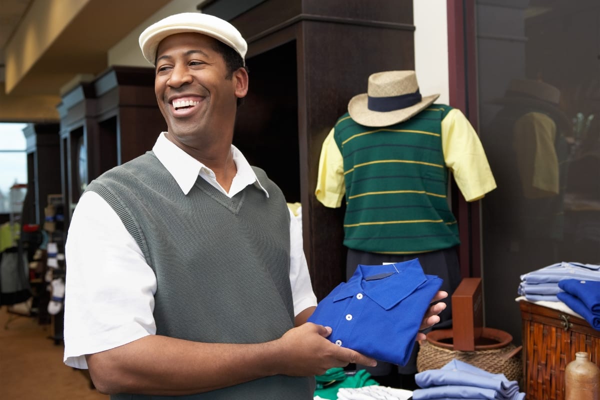 A person in a golf shop holds a folded blue polo shirt, smiling. Displayed behind are several shirts and a hat, all designed to enhance golf performance.