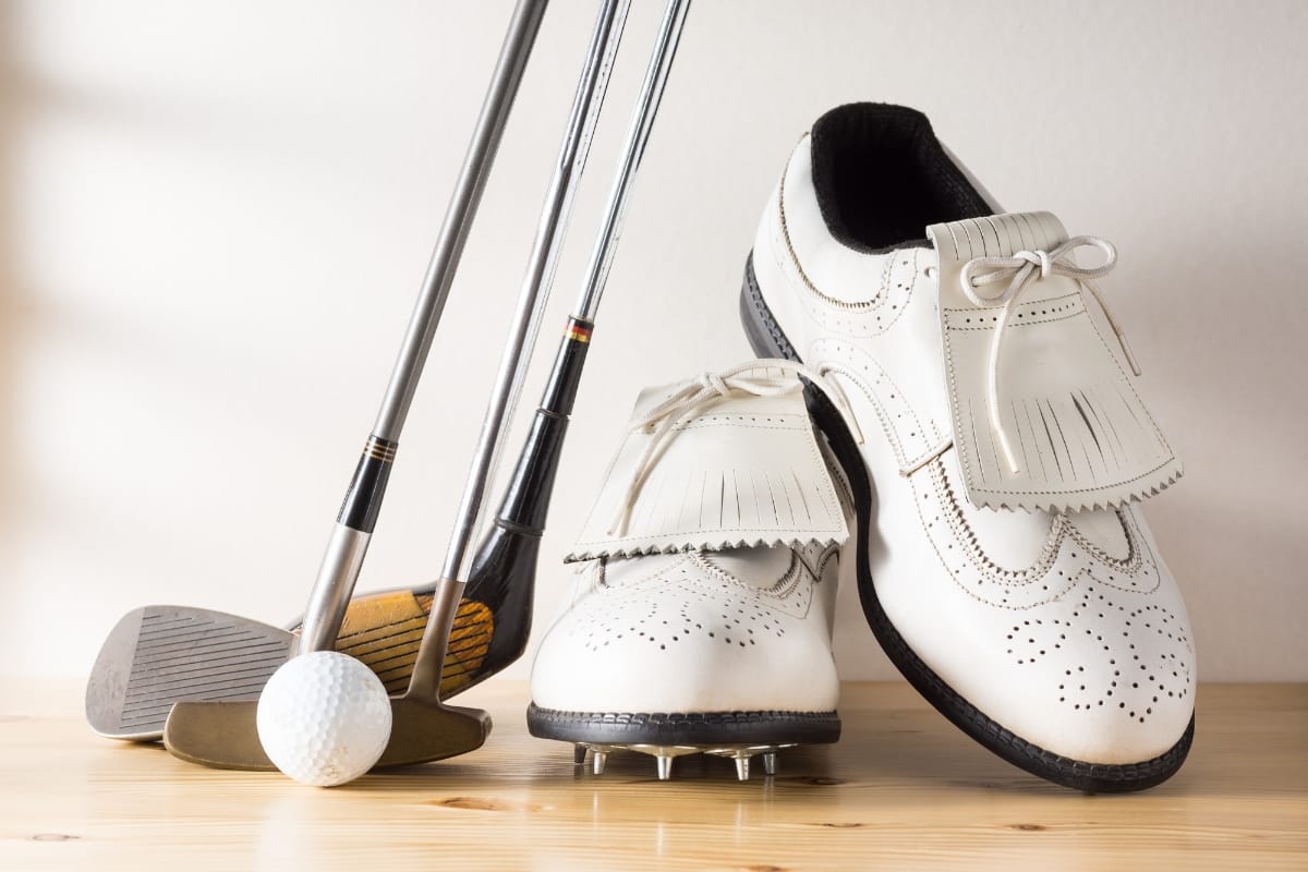 Golf clubs, a white golf ball, and a pair of performance-enhancing white golf shoes with spikes are arranged on a wooden surface.