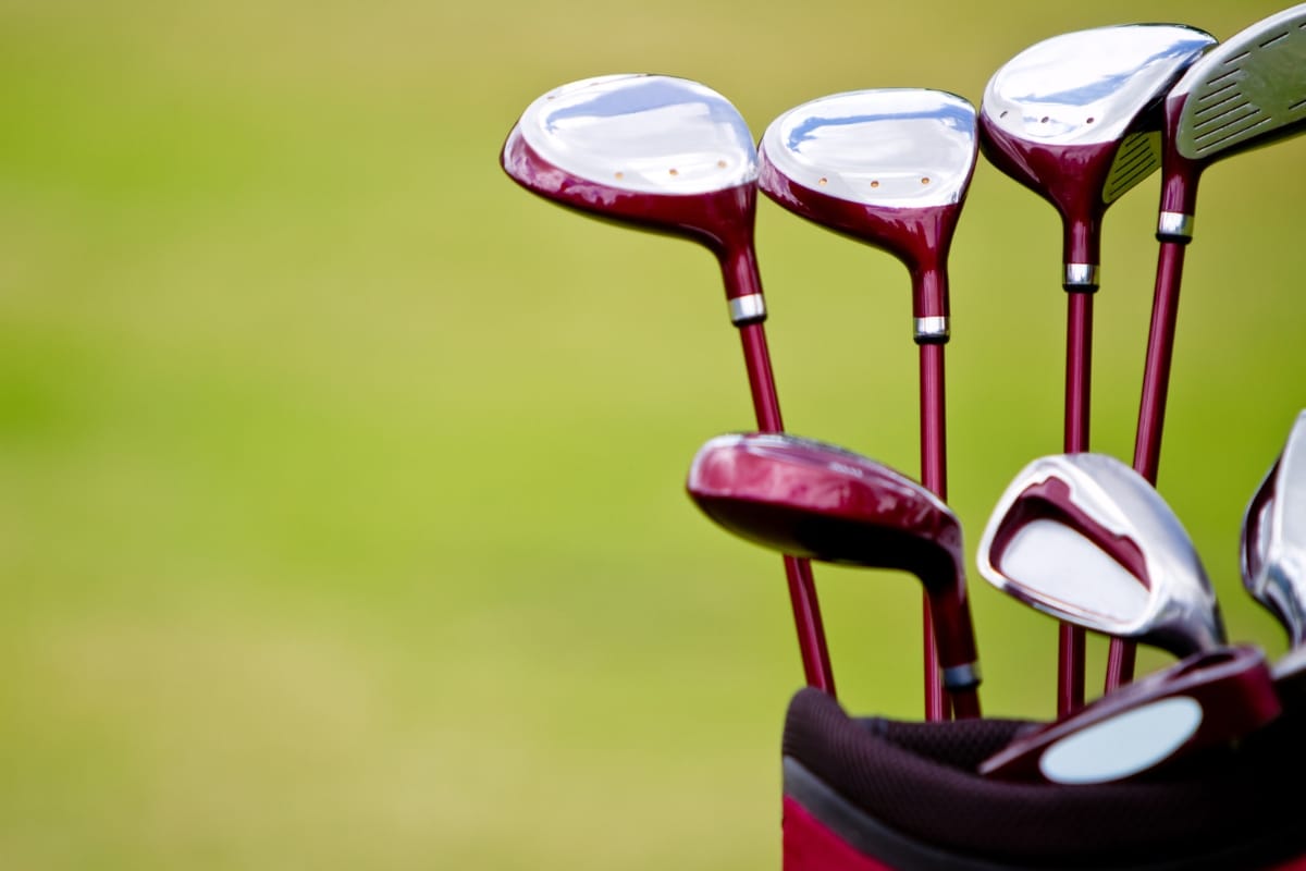 A set of golf clubs with sleek metallic heads and maroon shafts in a golf bag, crafted for optimum golf performance, set against a blurred green background.