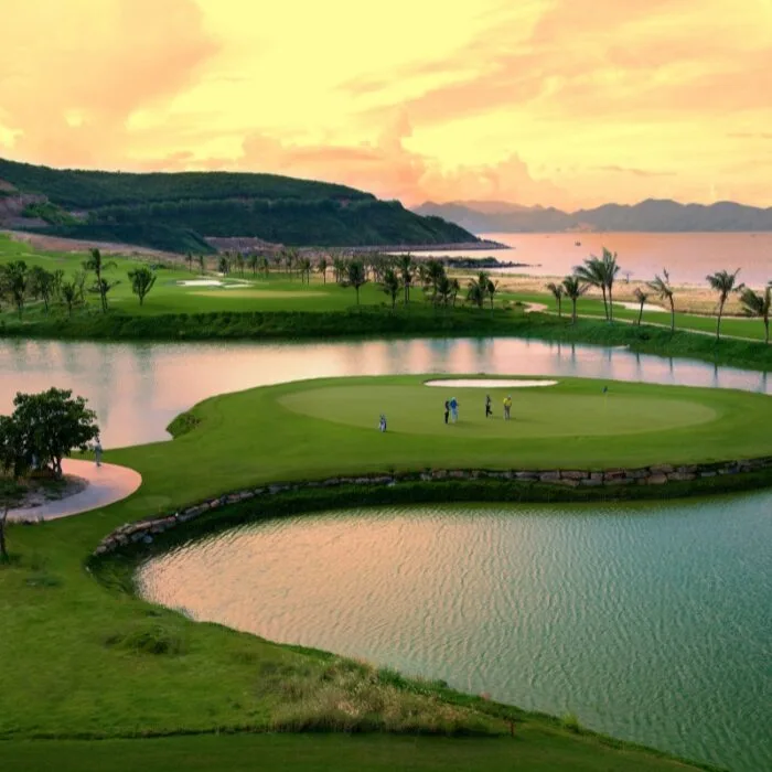 Aerial view of a sustainable golf course at sunset features an island green embraced by water, with a few people standing on the lush green. Hills and a serene body of water enhance the captivating backdrop.