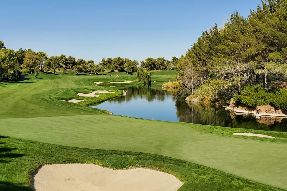 Sustainable golf course with lush green grass, sand bunkers, and a water hazard under a clear blue sky. Trees line the fairway.