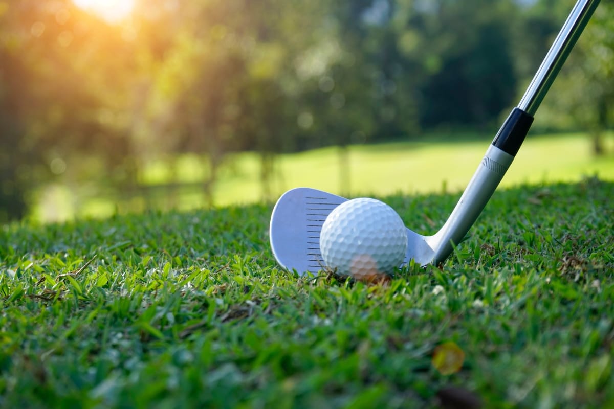 A golf club is positioned behind a golf ball on a sustainable course, with sunlight streaming through trees in the background.