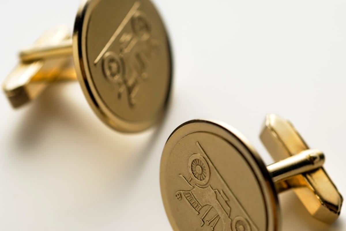 Two gold cufflinks with engraved designs of vintage cars rest on a white surface, adding a classic touch to any professional golf attire.