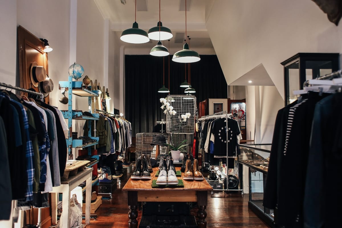 Boutique clothing store interior with racks of clothes featuring professional golf attire, displayed hats, and shoes on a central wooden table. Ceiling lights hang overhead, and a wire shelf holds white orchids in the background.