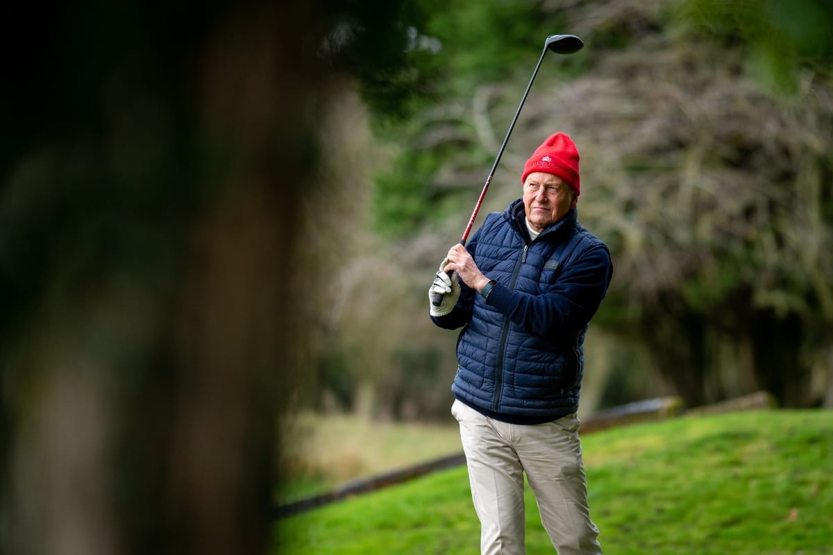 An older man in a red beanie and blue jacket, dressed in winter golf clothing, holds a golf club while standing on a grassy area with trees in the background.