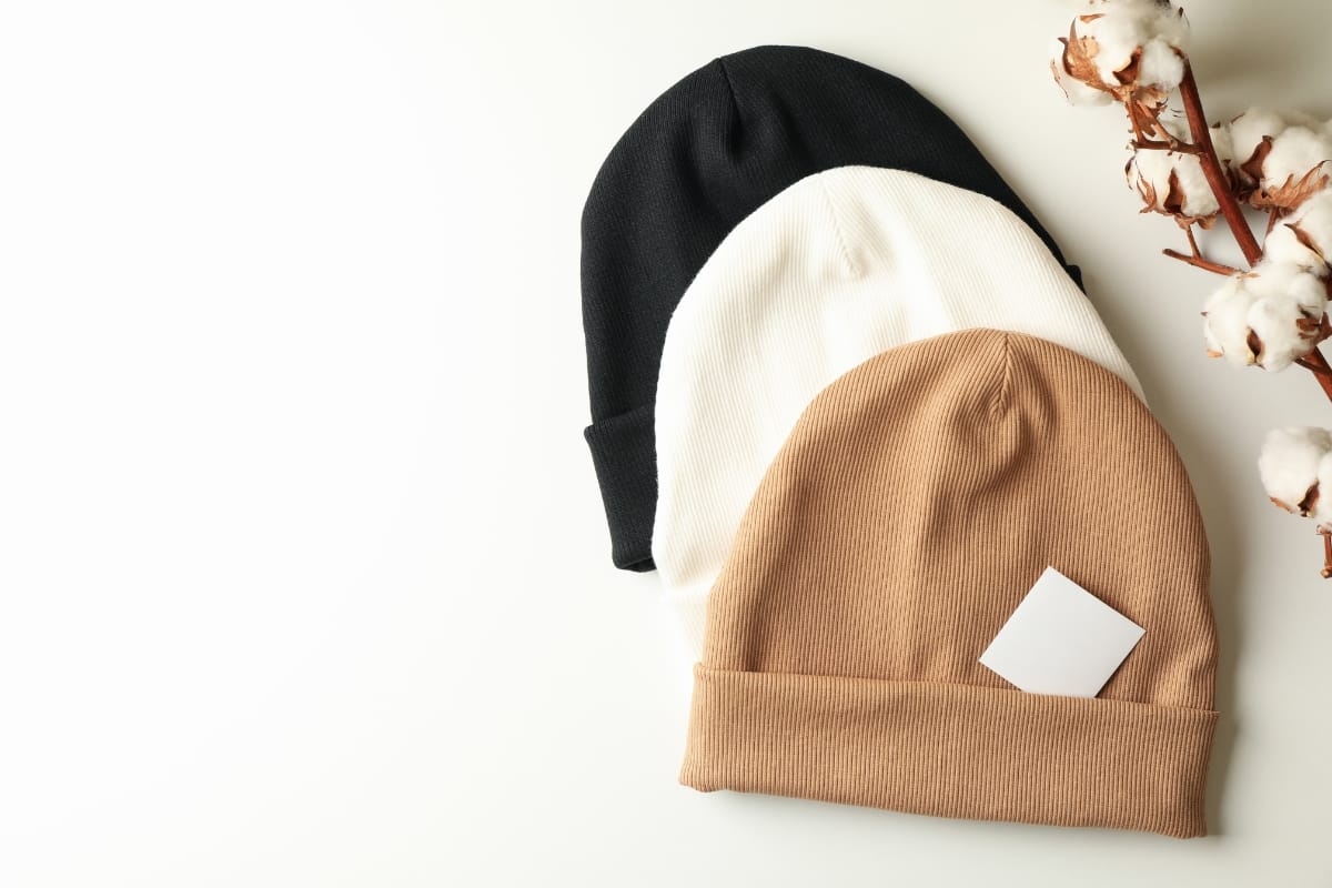 Three stylish knit beanies in black, white, and brown are stacked on a white surface next to cotton branches. A small blank card rests atop the top beanie, illustrating the benefits of wearing golf beanies with their versatile design and warmth for any chill day.