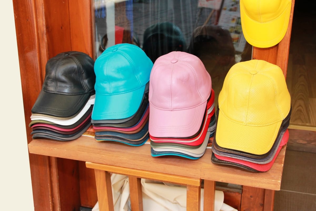 Stacks of leather baseball caps in various colors, including black, teal, pink, and yellow, displayed on wooden shelves.