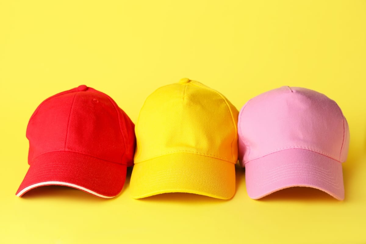 Three baseball caps in red, yellow, and pink are lined up against a solid yellow background.