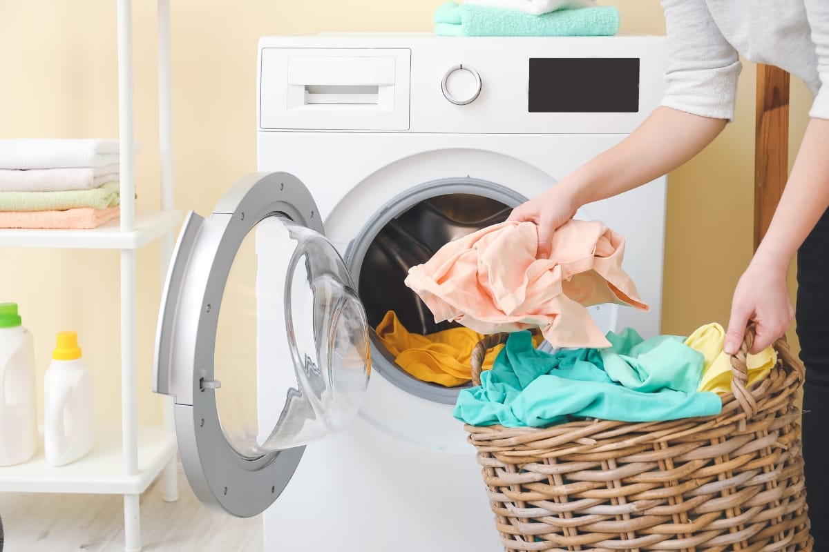 Person loading colorful clothes into a front-loading washing machine from a wicker basket, with detergent bottles on the side.