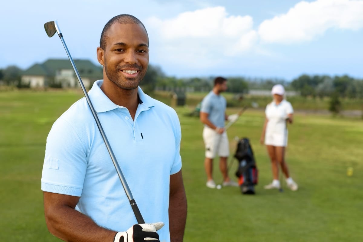 A man in a light blue polo shirt and the best golf shorts holds a golf club, smiling at the camera on a golf course, with two people and golf bags in the background.