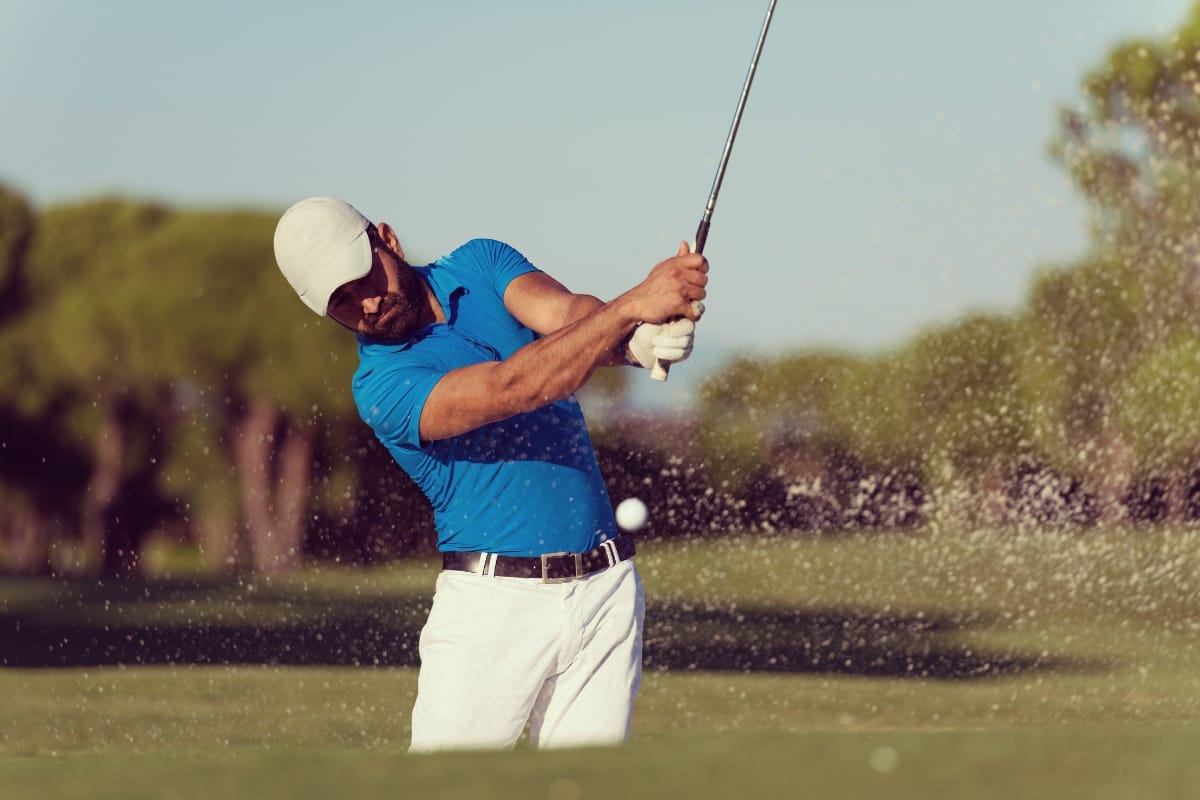 A man in a blue shirt, best golf shorts, and white cap hits a golf ball out of a sand bunker on a golf course, with sand flying in the air.