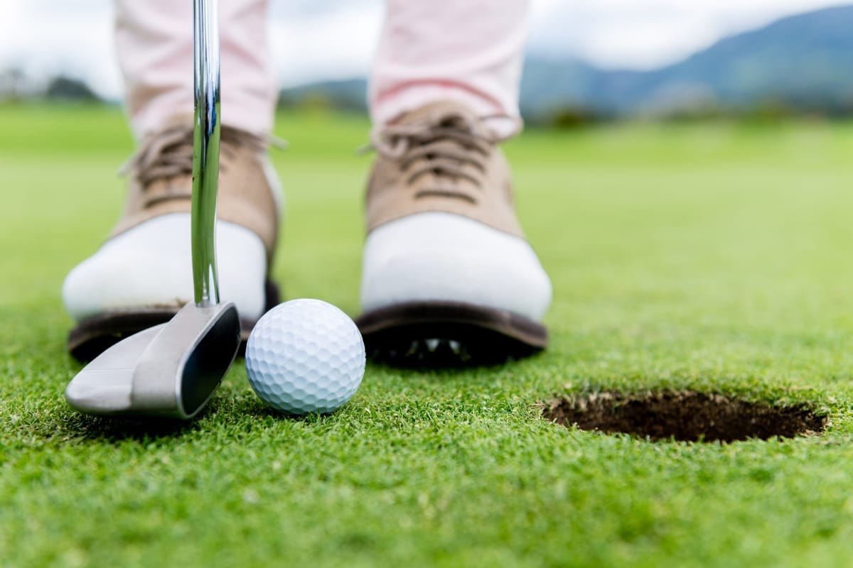 A person, wearing their Best Golf Shoes, prepares to putt a golf ball into the hole on a golf course, with a close-up view of the club, ball, and shoes.