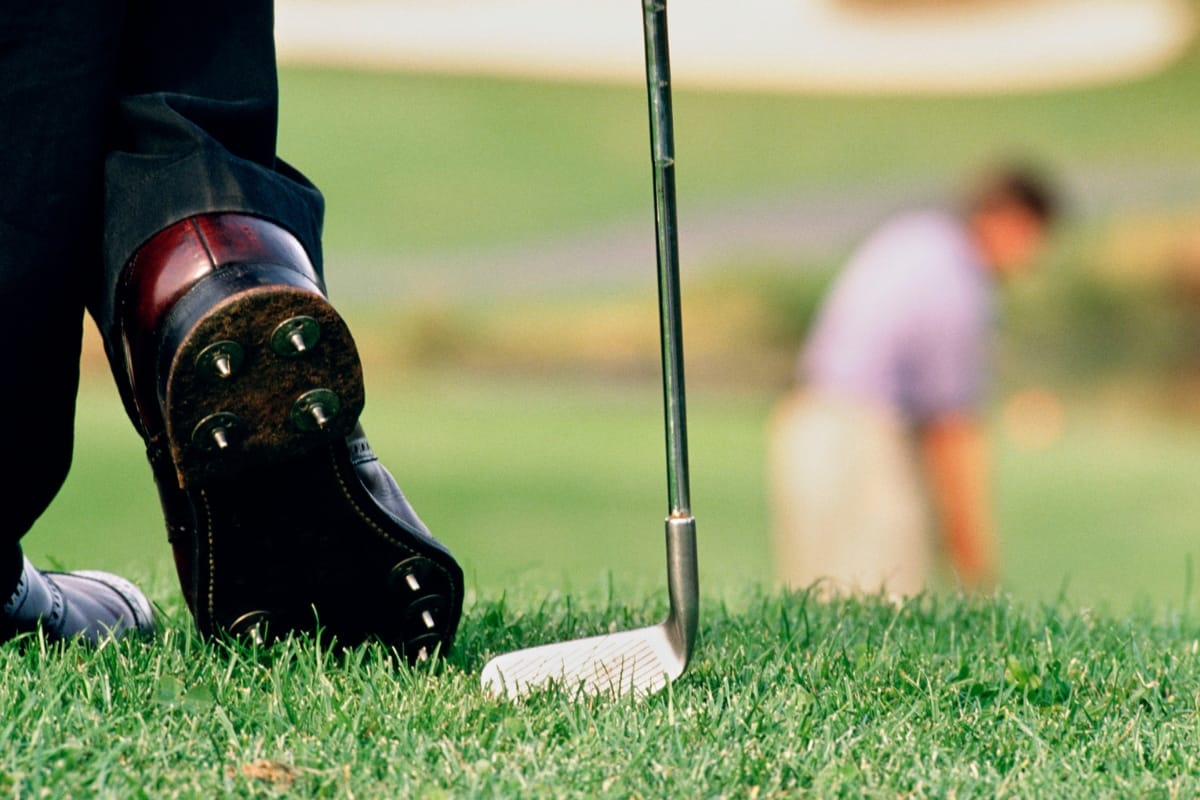 Close-up of a golfer's feet in the best golf shoes with a golf club on the grass, while another person stands blurred in the background on a golf course.