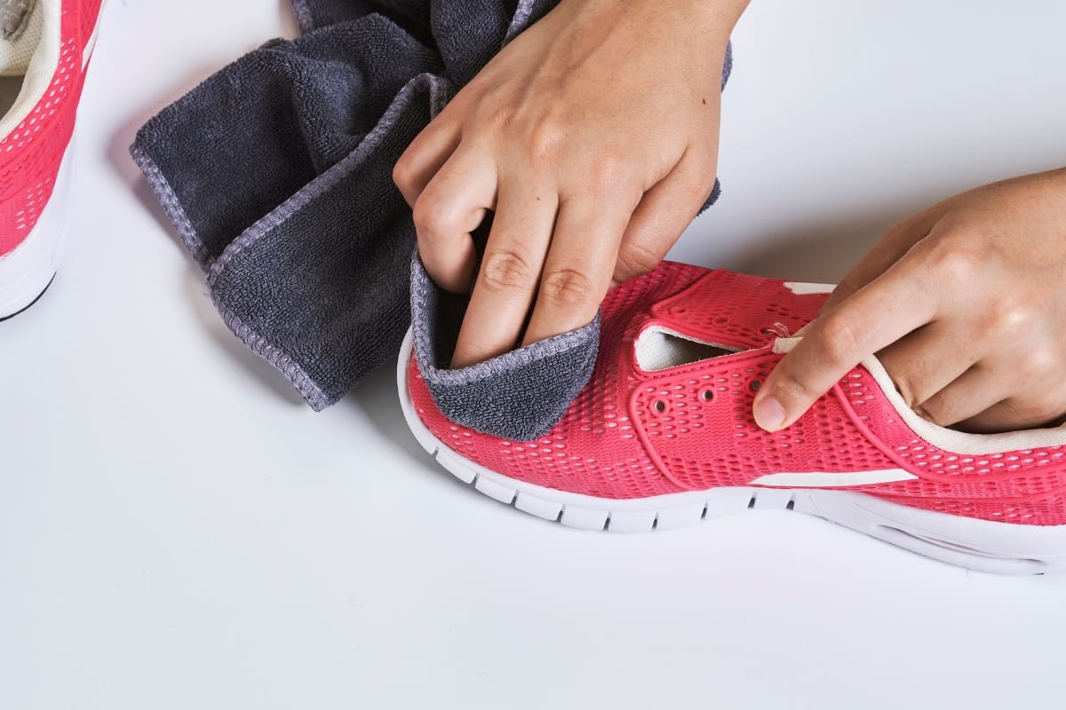 A person cleans a pink athletic shoe labeled as one of the Best Golf Shoes with a dark gray cloth on a white surface.