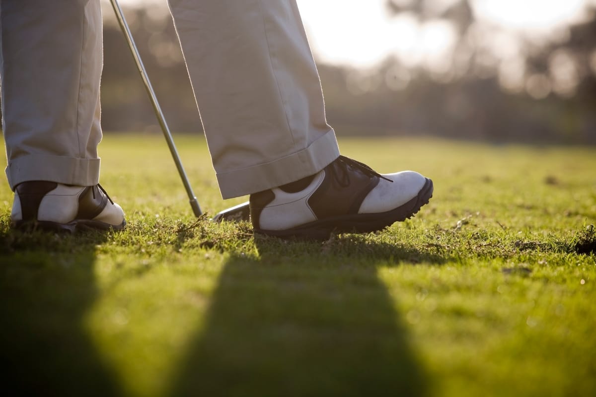 Close-up of a person wearing the best golf shoes and beige pants standing on a grassy golf course, holding a golf club.