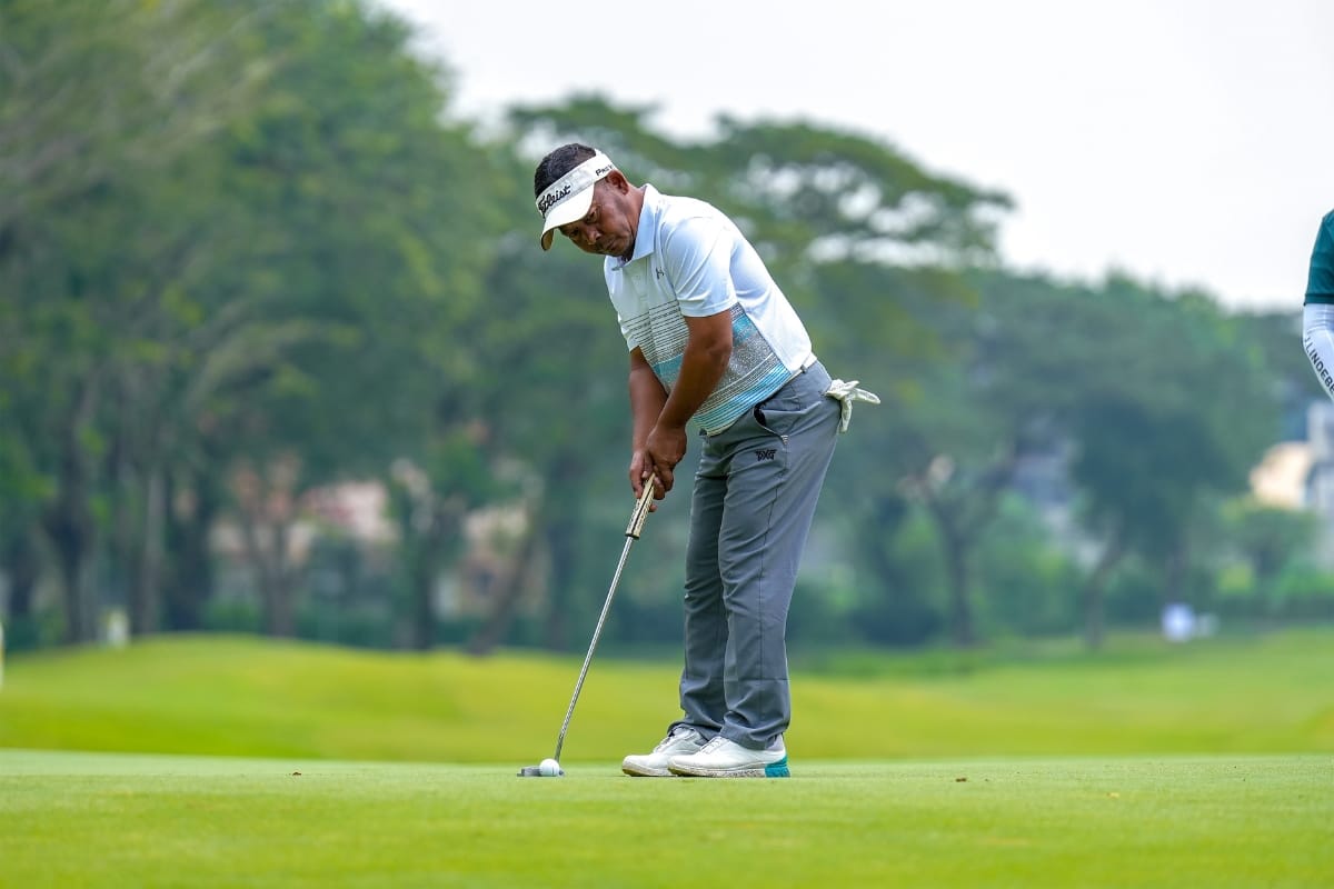 A golfer in a white polo and gray pants lines up a putt on a green golf course surrounded by trees, showcasing his precision and essential golf accessories.