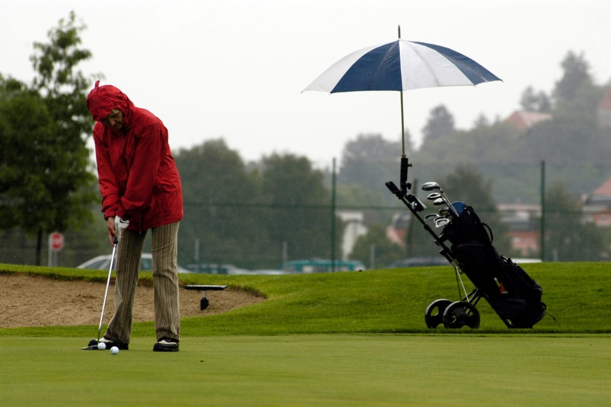A person in a red rain jacket putts on a golf green while Golf Accessories like an umbrella are attached to a golf bag and cart nearby on a cloudy day.