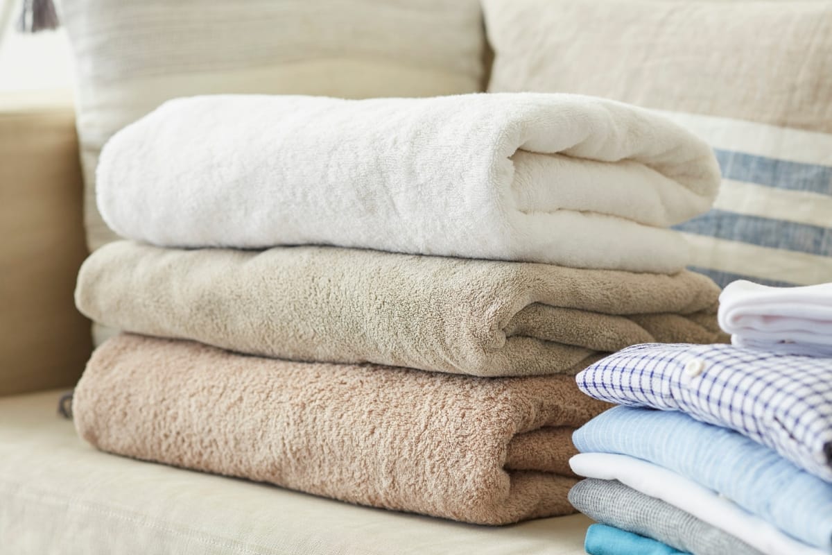 Three folded towels in white, beige, and tan are stacked on a couch next to a pile of folded shirts with different patterns and colors, alongside some golf accessories for a touch of sporty elegance.