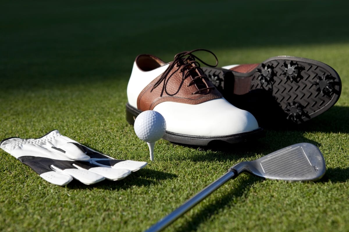 Golf accessories such as golf shoes, a glove, a golf ball on a tee, and an iron club are arranged on green grass.