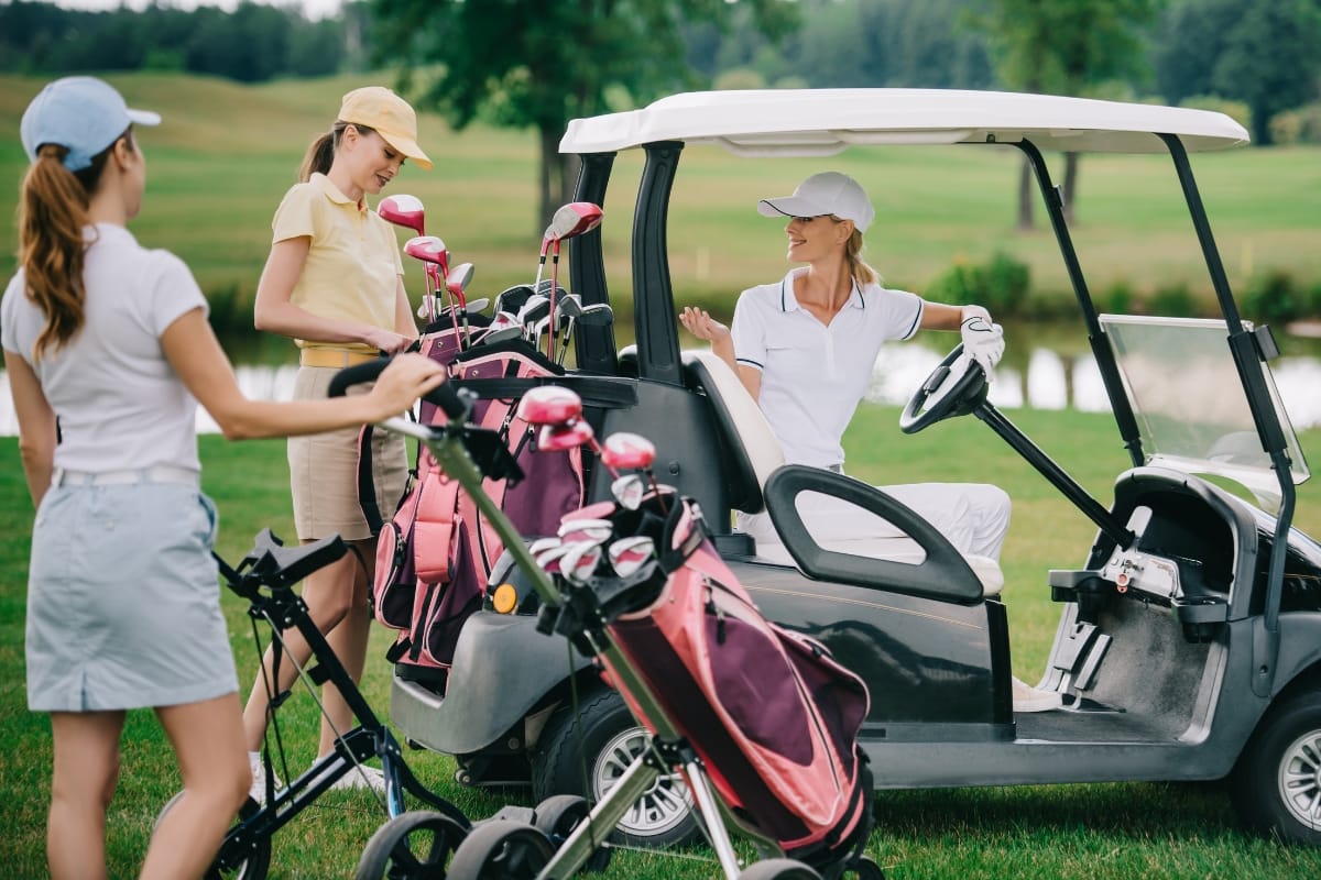 Three women in stylish golf apparel with clubs stand by a golf cart on a grassy course, chatting and preparing to play.