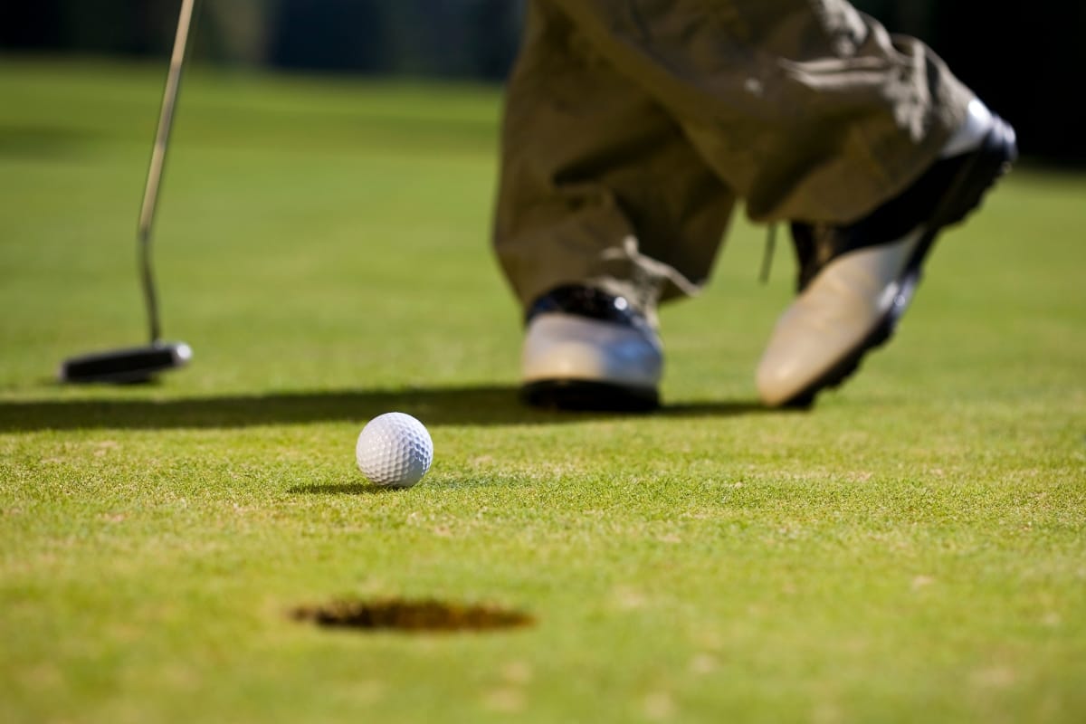 Close-up of a golf ball on the green near a hole, with a golfer in the background preparing to putt in their stylish golf socks.