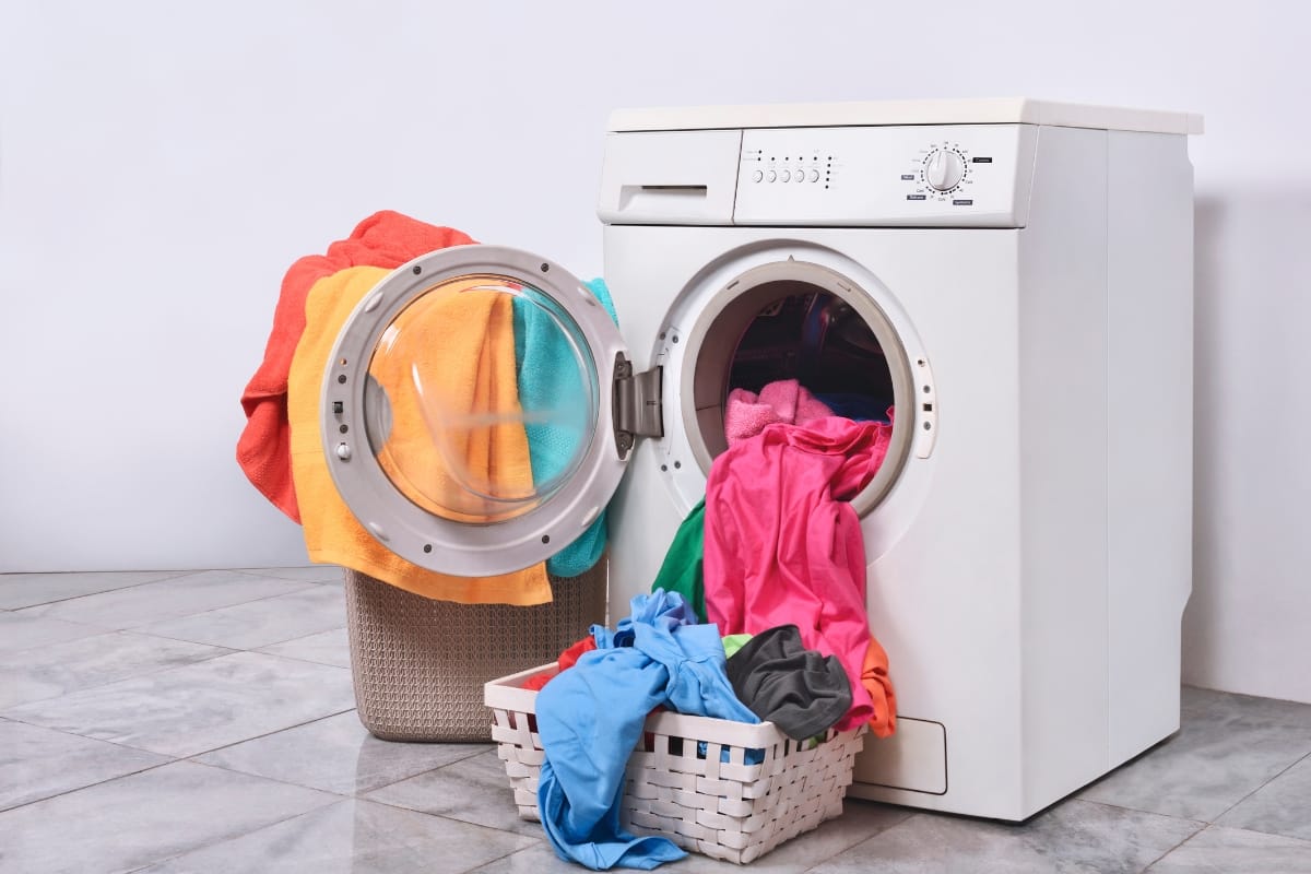 A front-loading washing machine with its door open is surrounded by colorful laundry, including moisture-wicking golf shirts, spilling from baskets onto the tiled floor.