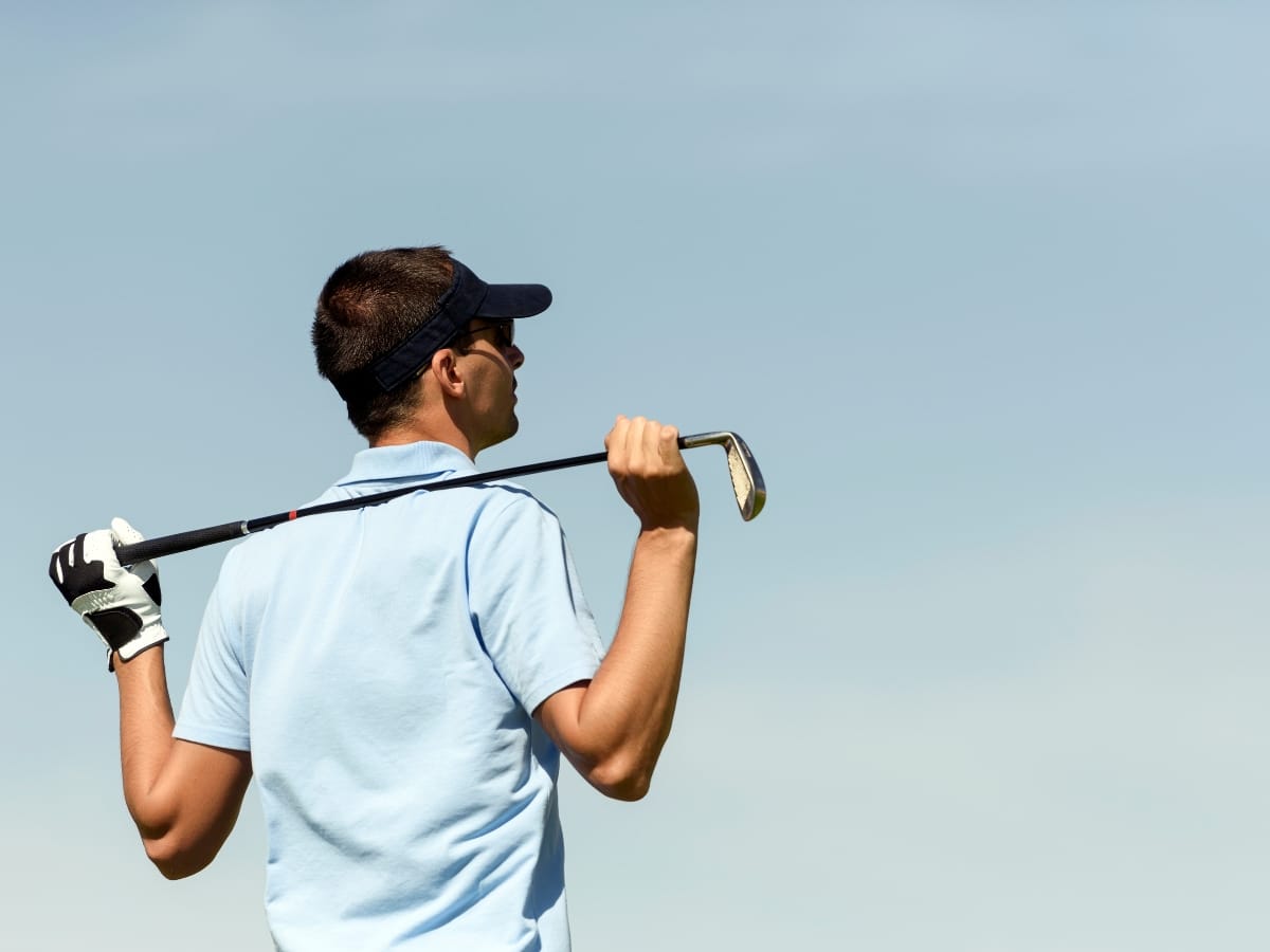A man wearing one of the best golf visors and a golf glove holds a club over his shoulders while looking into the distance against a clear sky.