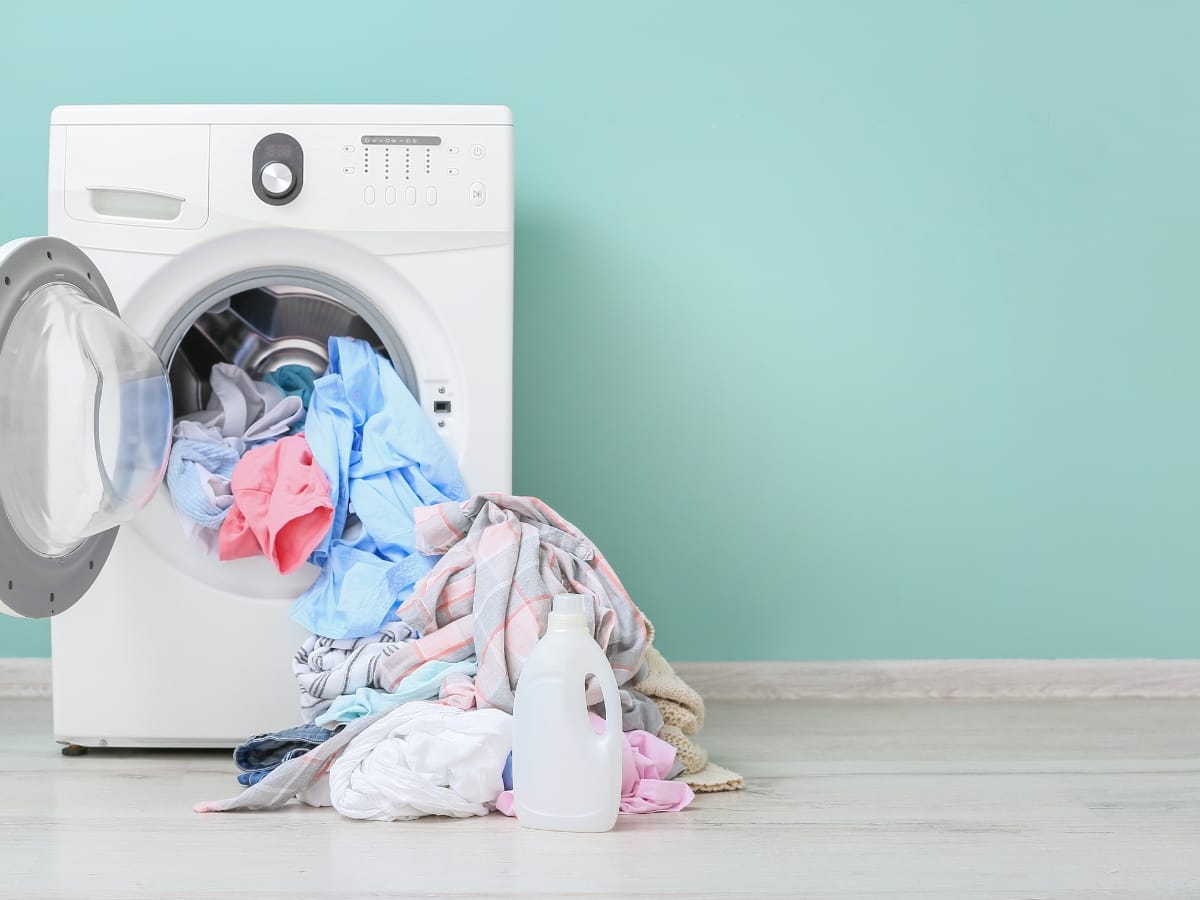 A front-loading washing machine with its door open has laundry spilling out onto the floor. Nearby, a bottle of detergent and one of the best golf visors rest against a light blue wall.