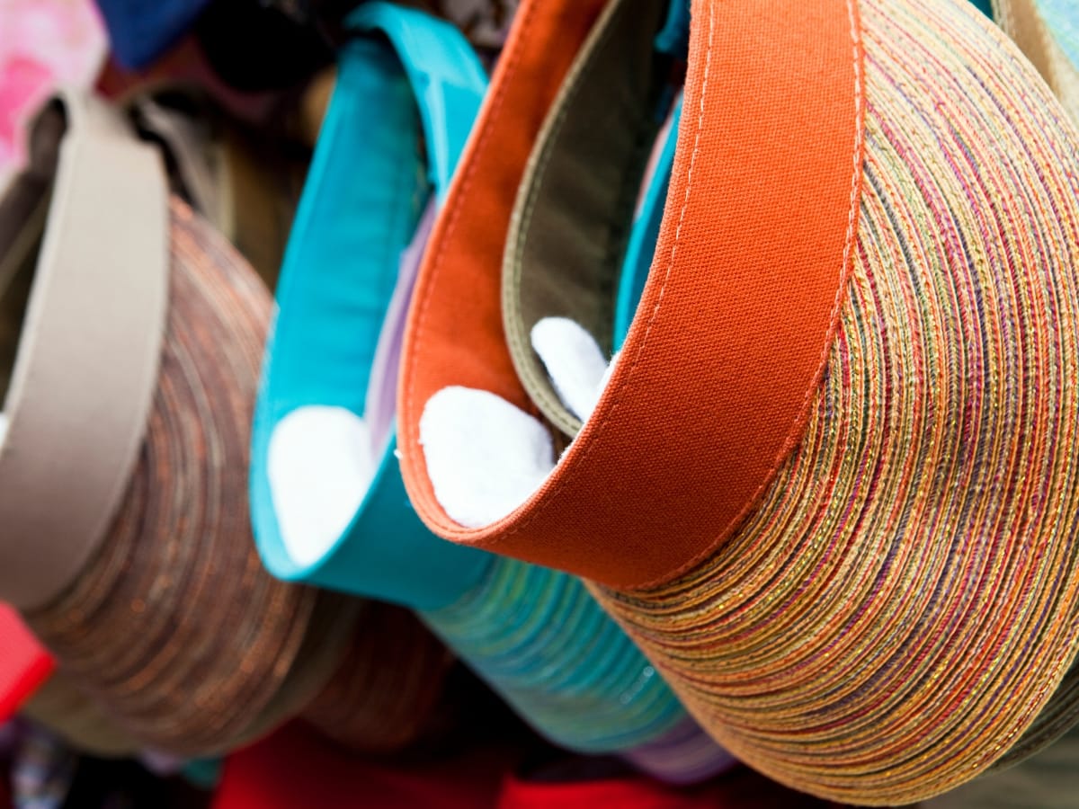 Several colorful sun visors made of woven material are displayed hanging in a row, their textured brims and fabric bands offering both style and sun protection—perfect picks for those seeking the best golf visors.