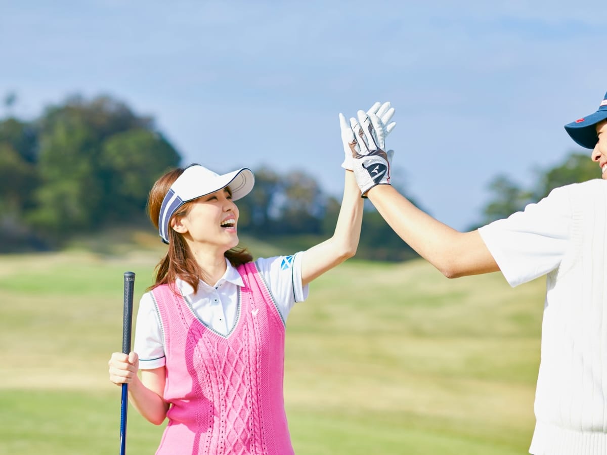 Two people wearing golf attire and the best golf visors high-five on a golf course, with one holding a golf club. The background shows grass and trees under a clear sky.