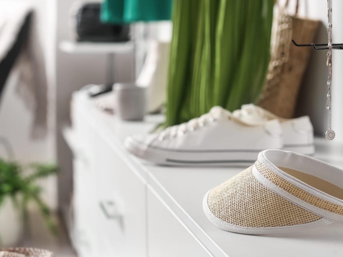 A beige mesh visor, among the best golf visors, and a pair of white sneakers rest on a white dresser in a brightly lit room with clothes and accessories in the background.