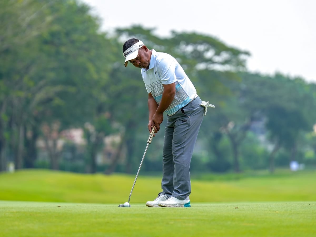 A golfer in a light blue shirt, gray pants, and one of the best golf visors is putting on a green golf course with trees in the background.