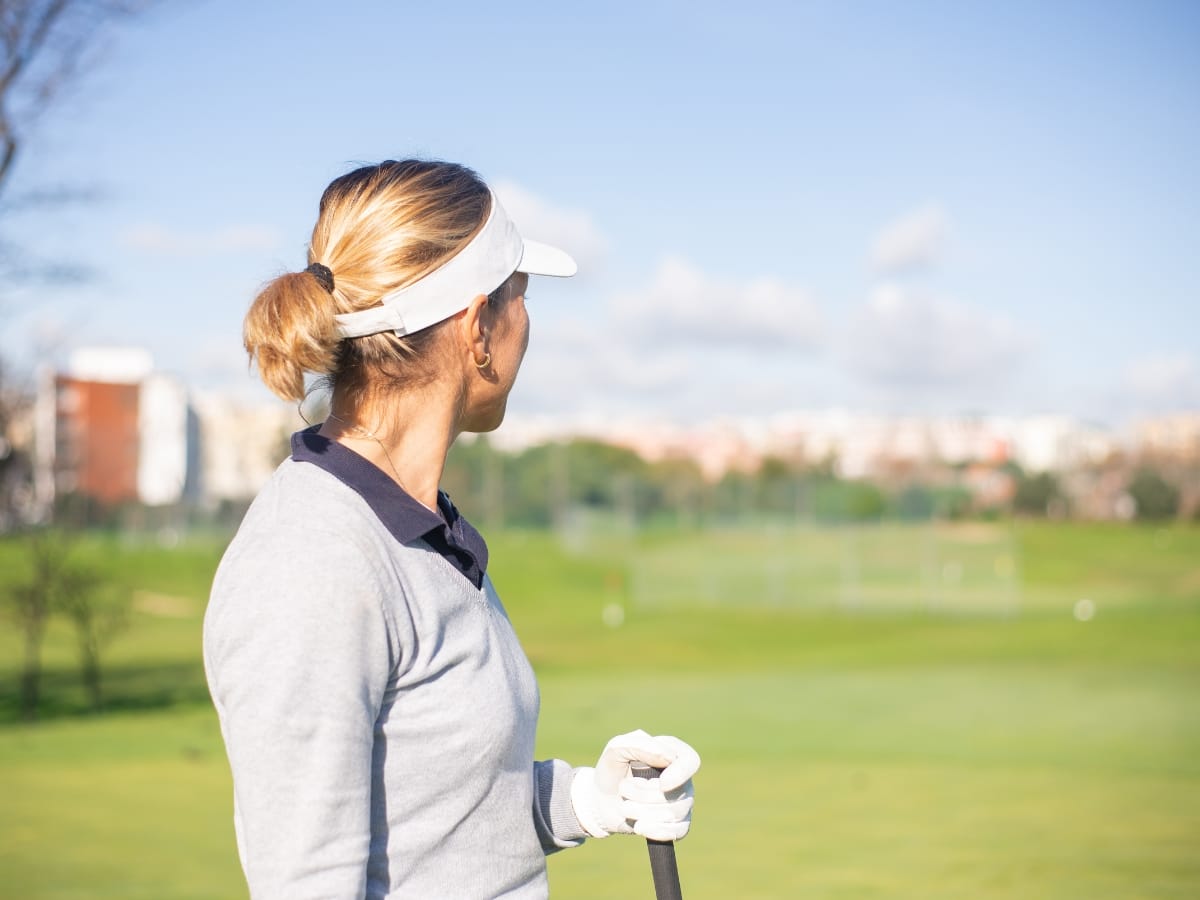 A woman wearing one of the best golf visors and a glove stands on a golf course, holding a club and looking into the distance on a sunny day.