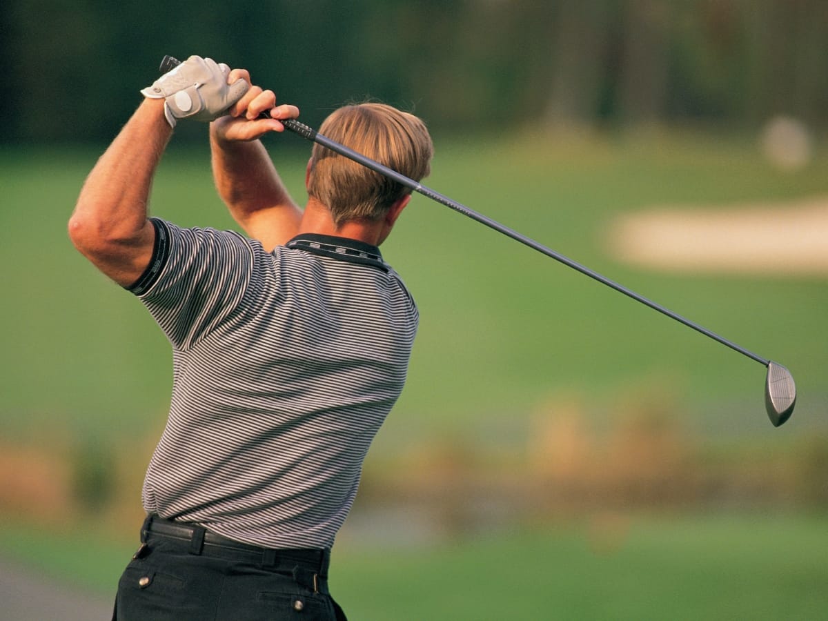 A person wearing a striped shirt, a classic choice when considering what to wear for a golf tournament, swings a golf club on a golf course, facing away from the camera.