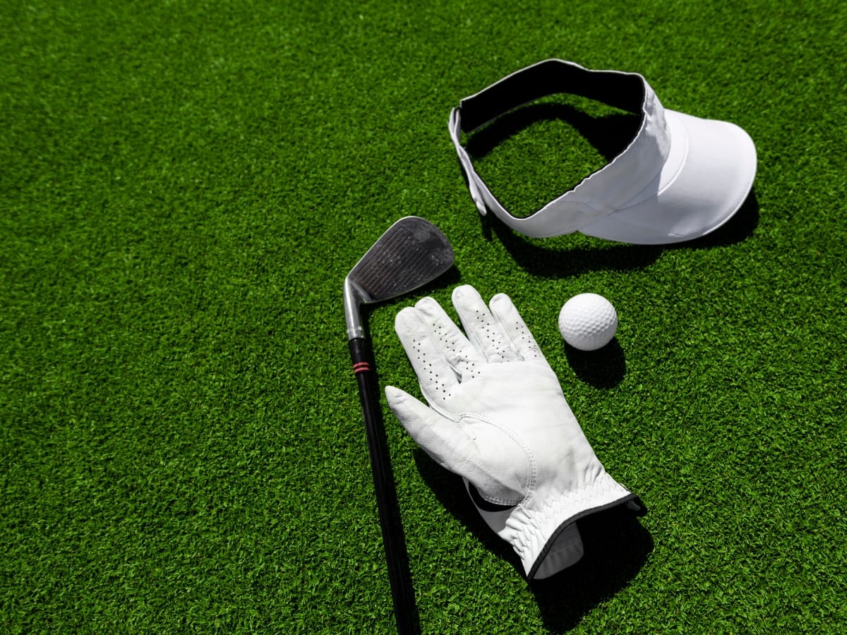 A golf club, white glove, golf ball, and white visor—essentials when considering what to wear for a golf tournament—are placed on green artificial turf.