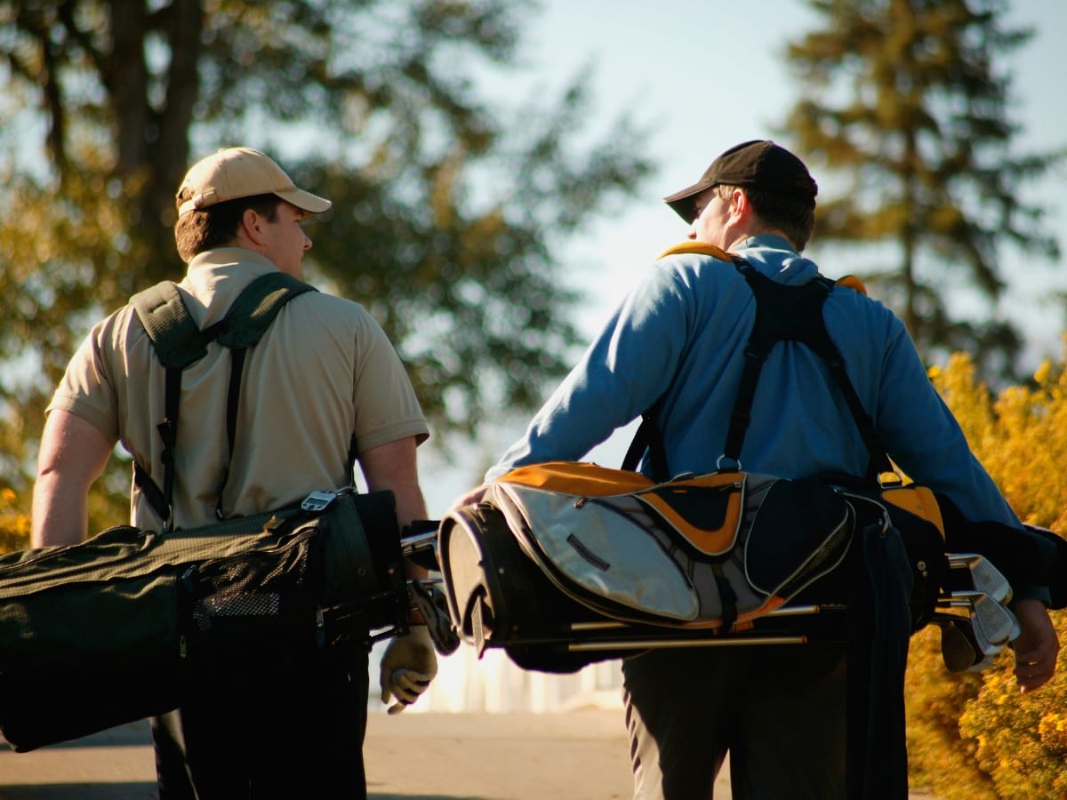 Two people walk side by side, each carrying a golf bag over their shoulders, dressed smartly for the course—an inspiring look at what to wear for a golf tournament on a sunny day with trees in the background.