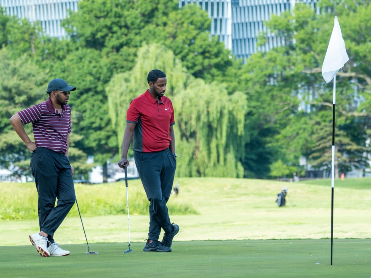 Two men stand on the golf green near the hole, both holding clubs and dressed in outfits perfect for what to wear for a golf tournament, with trees and a building in the background.