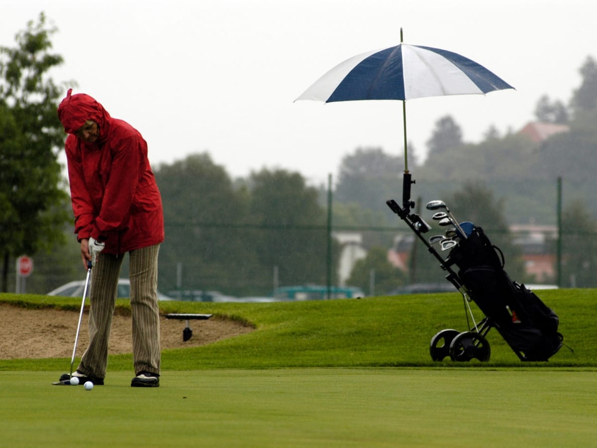 A person in a red jacket, equipped with the best golf rain gear, putts a ball on a wet green, with a golf bag and umbrella set on a trolley nearby.