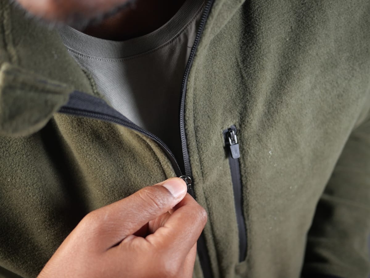 A person’s hand is shown zipping up a green fleece jacket, highlighting the zipper and fabric texture—perfect for mastering how to layer golf clothing for fall weather.