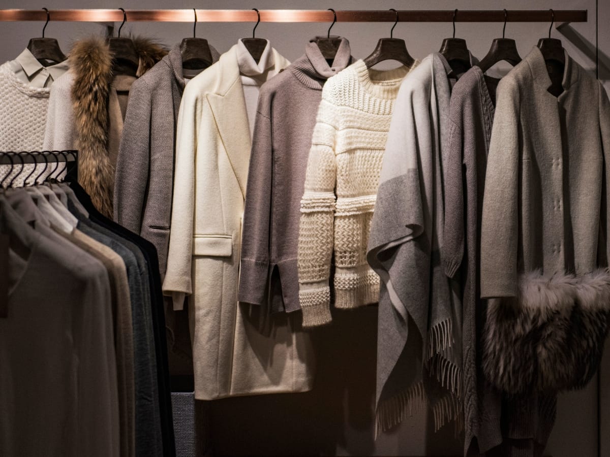 A row of neutral-colored coats, sweaters, and shawls on hangers in a wardrobe showcases various textures—perfect inspiration for how to layer golf clothing for fall weather with knits, wool, and fur accents.