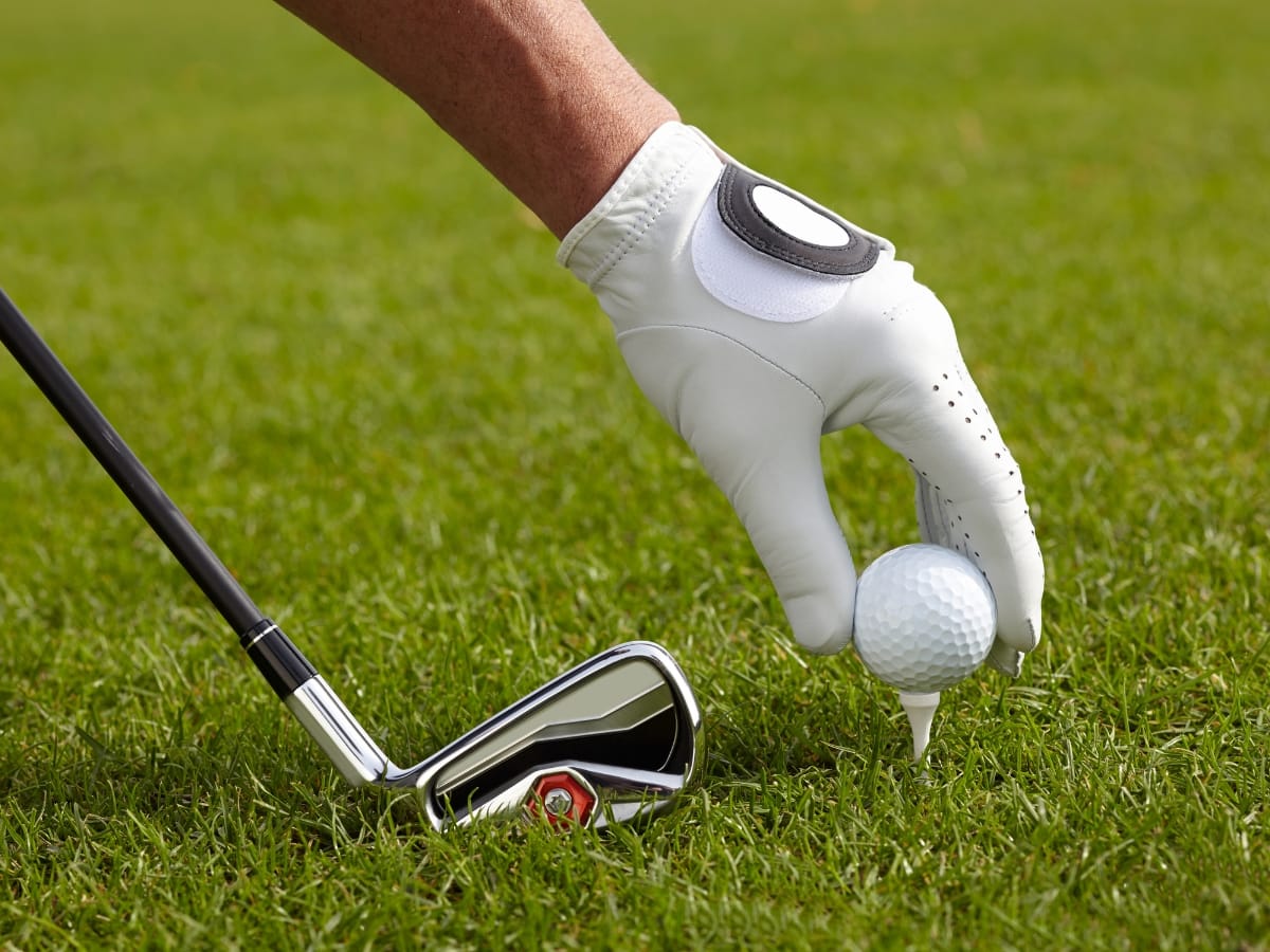 A gloved hand places a golf ball on a tee next to an iron golf club on grass, demonstrating the focus and preparation essential for the game—including knowing how to layer golf clothing for fall weather.