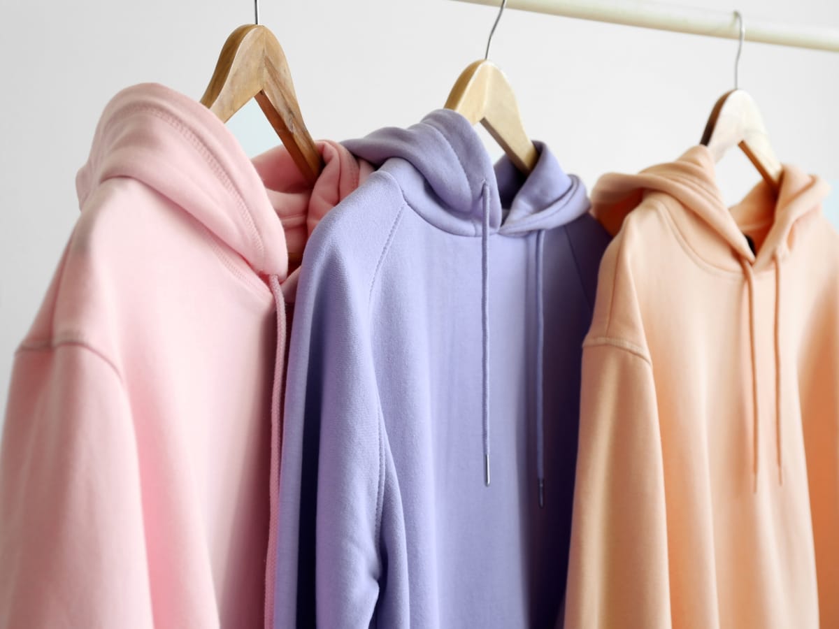 Three hooded sweatshirts in pastel pink, lavender, and beige—perfect as the best golf hoodies—hang on wooden hangers on a white clothing rack against a plain background.