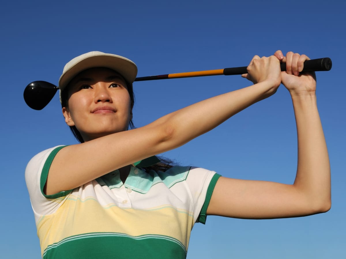 A person showcasing golf fashion for women, wearing a cap and holding a golf club in a follow-through pose against a clear blue sky.