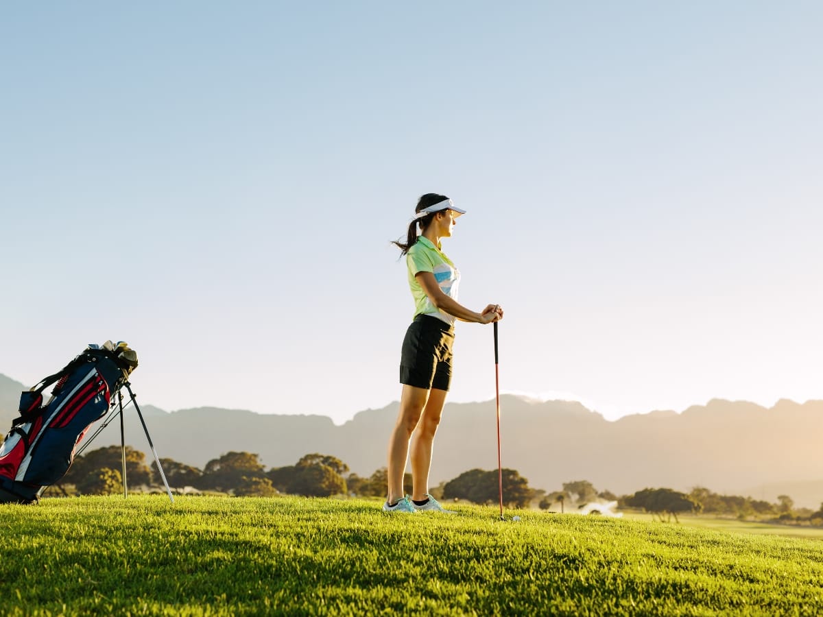 Embracing golf fashion for women, a woman in a chic visor and shorts stands on a golf course at sunset, holding a golf club, with her stylish golf bag nearby.