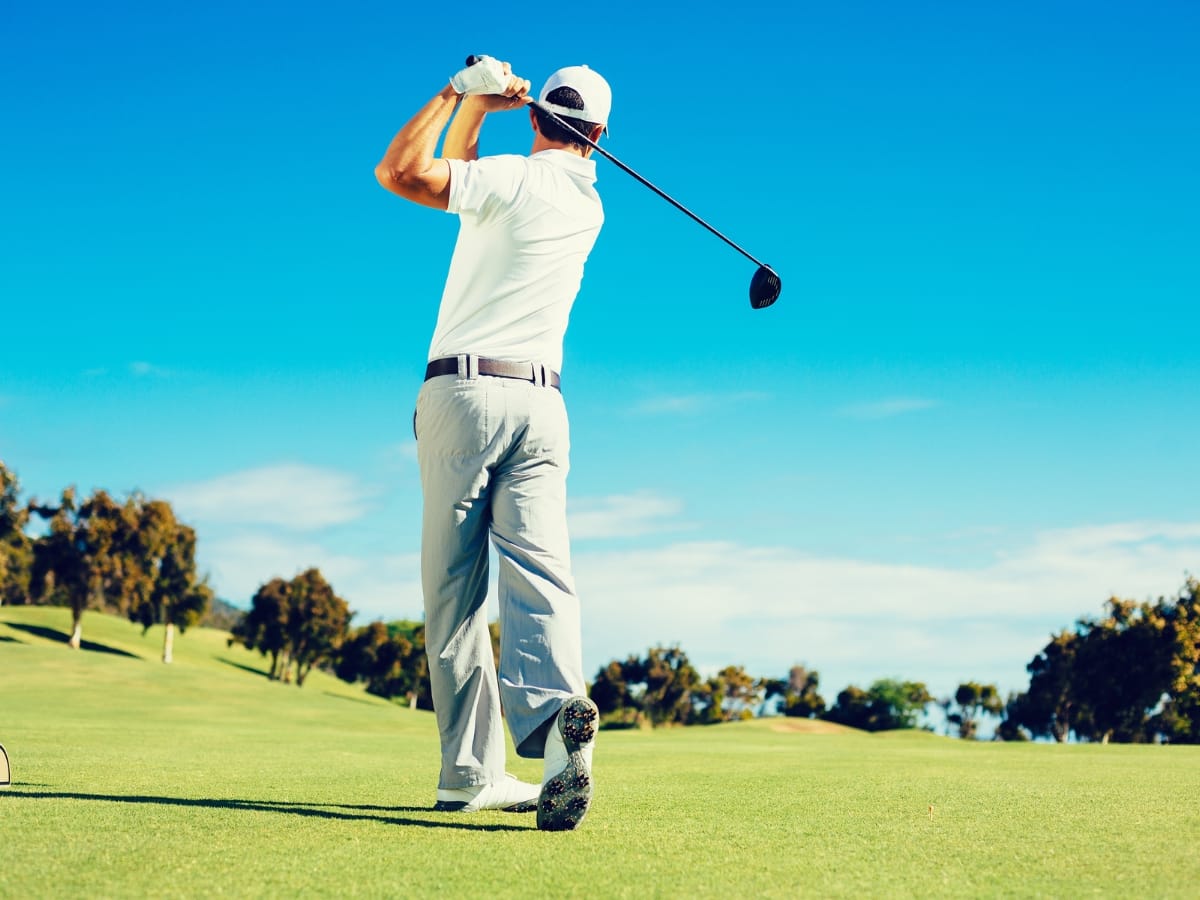 A man in white golf attire swings a club on a sunny course, surrounded by green grass and trees—perfect inspiration for Golf Gift Ideas for the Holidays.