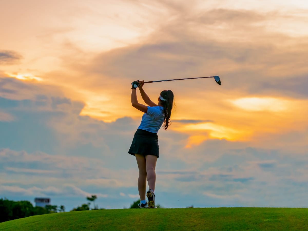 A woman swings a golf club on a grassy hill at sunset, with dramatic clouds and orange light in the background—capturing inspiration for Golf Gift Ideas for the Holidays.