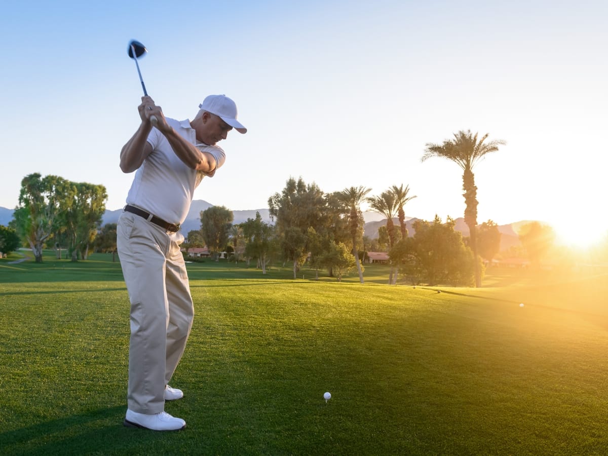 A man in a white shirt and cap, using the best golf gear for the New Year, prepares to swing a golf club on a sunny golf course at sunset, with trees and mountains in the background.