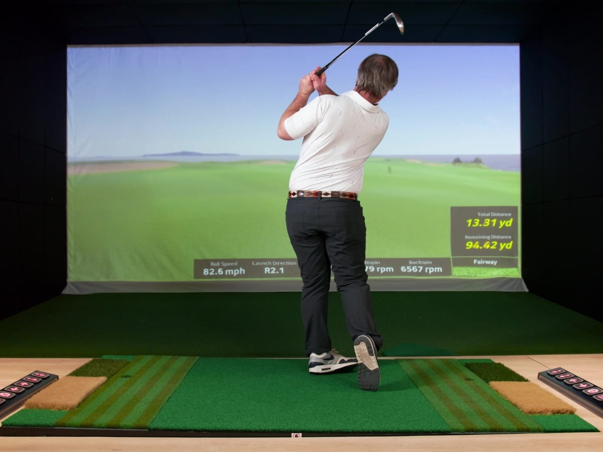 A man swings a golf club in an indoor golf simulator, watching the virtual course and shot statistics displayed on a large screen as he tests out the Best Golf Gear for the New Year.