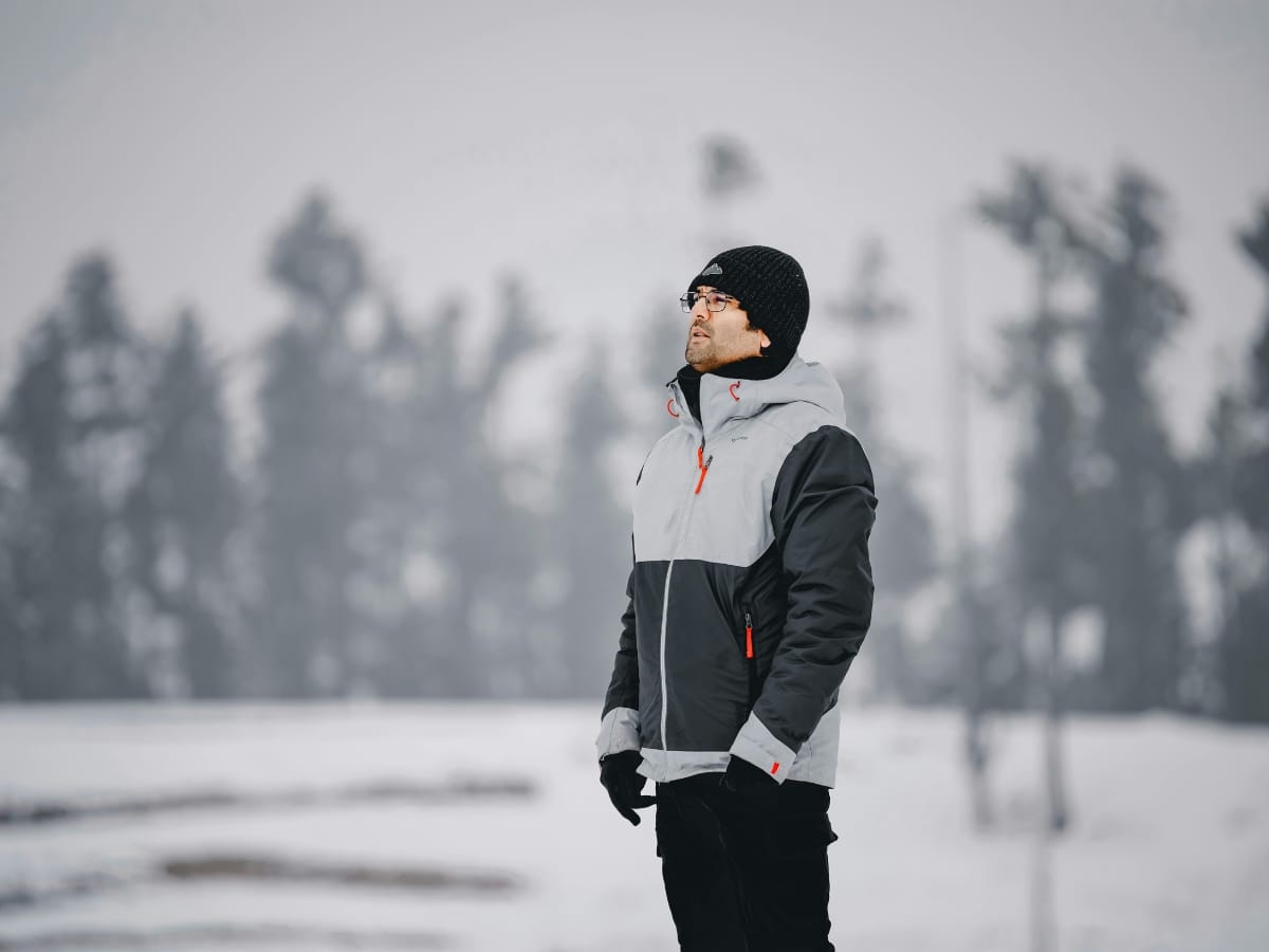 A person dressed in winter golf apparel—including a jacket, hat, and gloves—stands outdoors in a snowy landscape with blurred trees in the background.